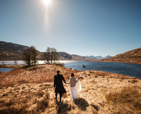 Bride and groom walking down to the shores of loch arklet in the sunshine after their wedding ceremony in the Trossachs, Scotland.